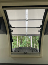 Inside an RV rental, an open window with a pull-down shade reveals lush green trees and a diamond plate surface.