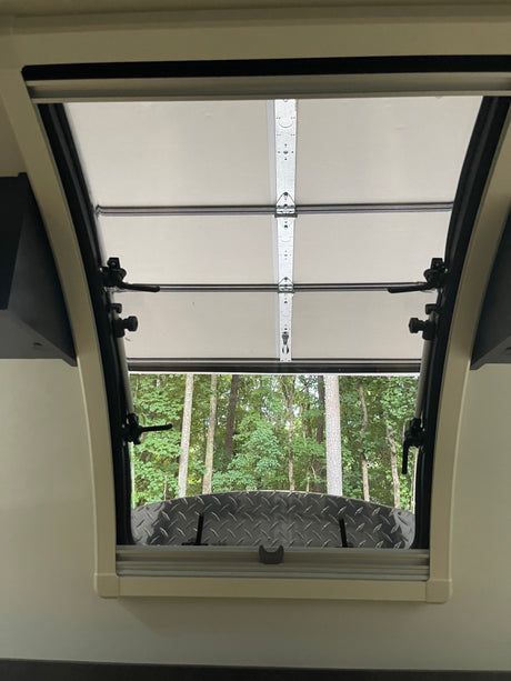 Inside an RV rental, an open window with a pull-down shade reveals lush green trees and a diamond plate surface.