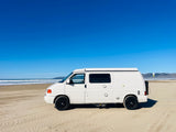 A white RV rental van sits on a sandy beach with ocean waves and clear blue sky, perfect for outdoor travel.