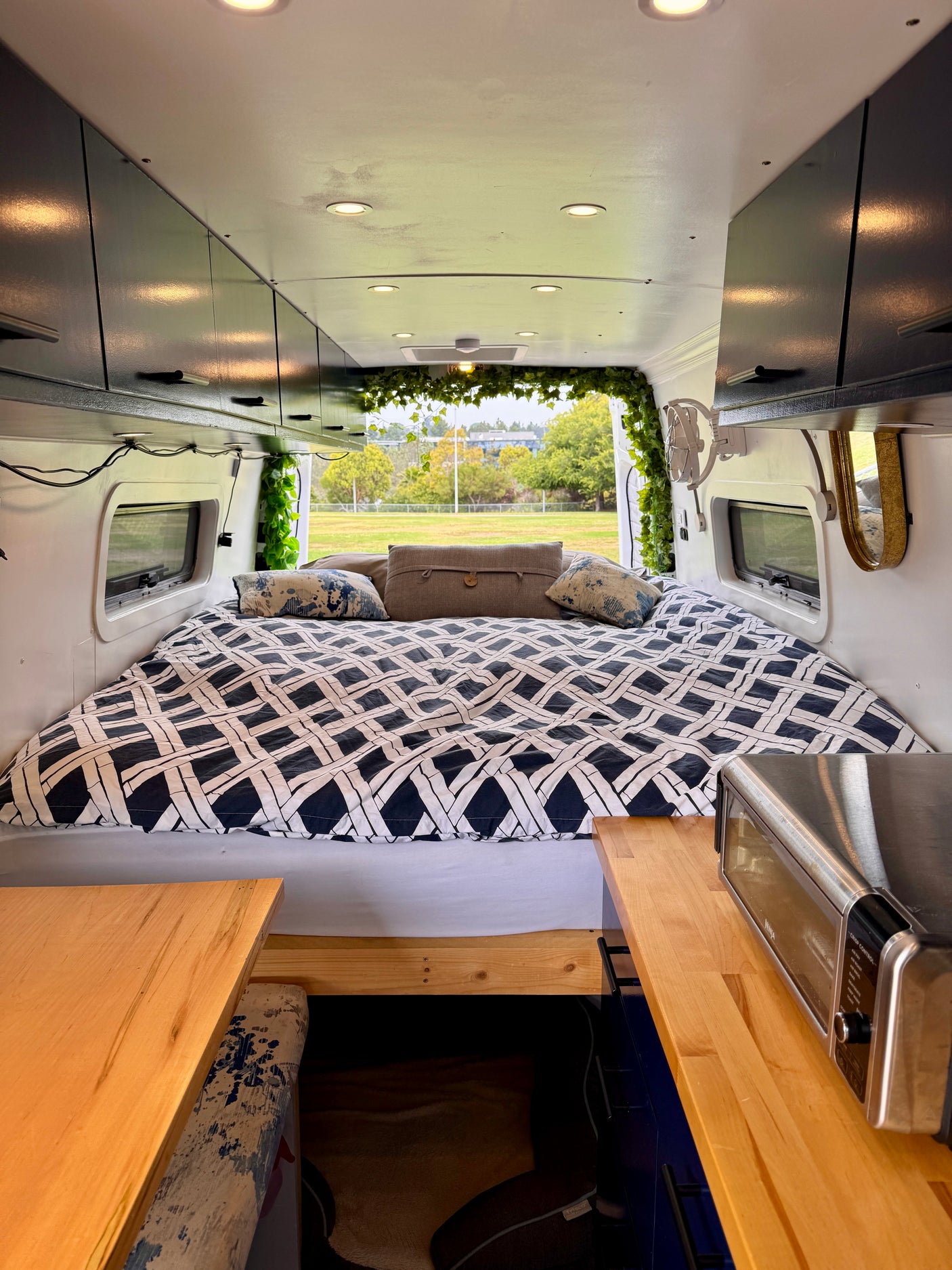 Modern RV rental interior: cozy bed, overhead cabinets, kitchen area with toaster, and leafy green decor. Ready for adventure!