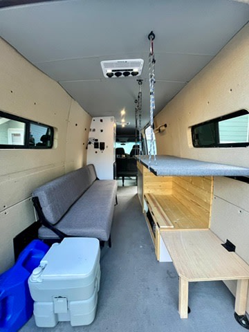 Explore this RV rental's spacious camper van interior, showcasing a long bench, portable toilet, and pull-out table/counter.