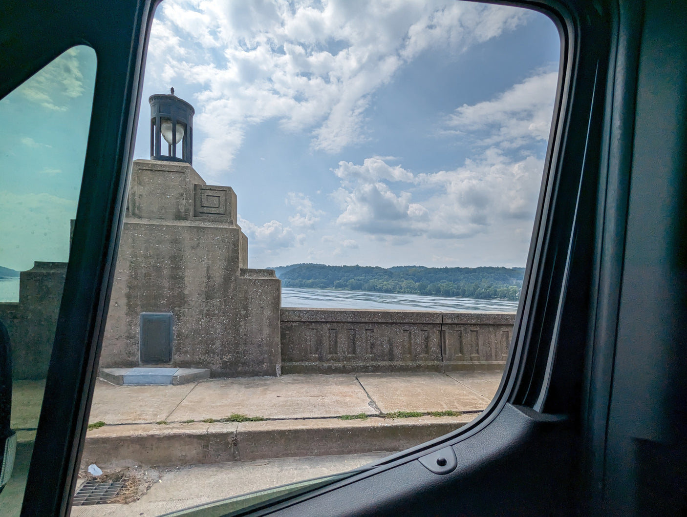 Scenic river view from an RV rental window, showcasing a concrete structure with a large lamp and distant forested hills.