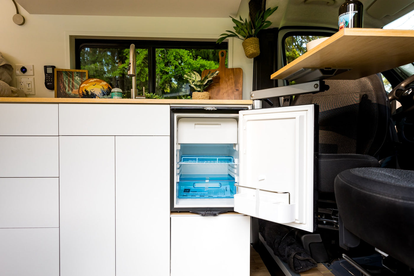 Bright rv rental van kitchen: compact design with open fridge, sink, wood counter, and green forest views.