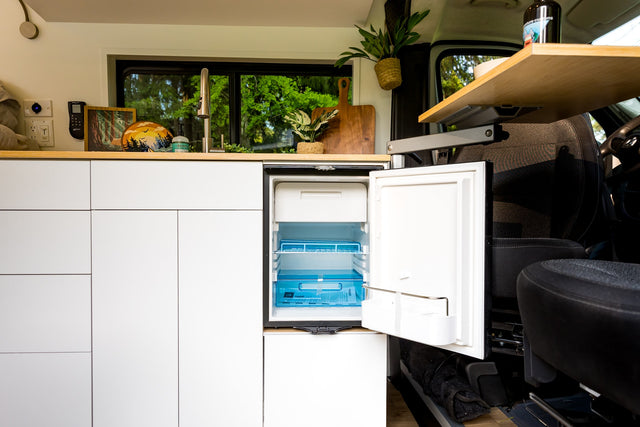 Bright rv rental van kitchen: compact design with open fridge, sink, wood counter, and green forest views.