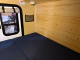 RV rental sleeping area: a dark blue mattress, light wood paneling, and a window with a view of a boat.
