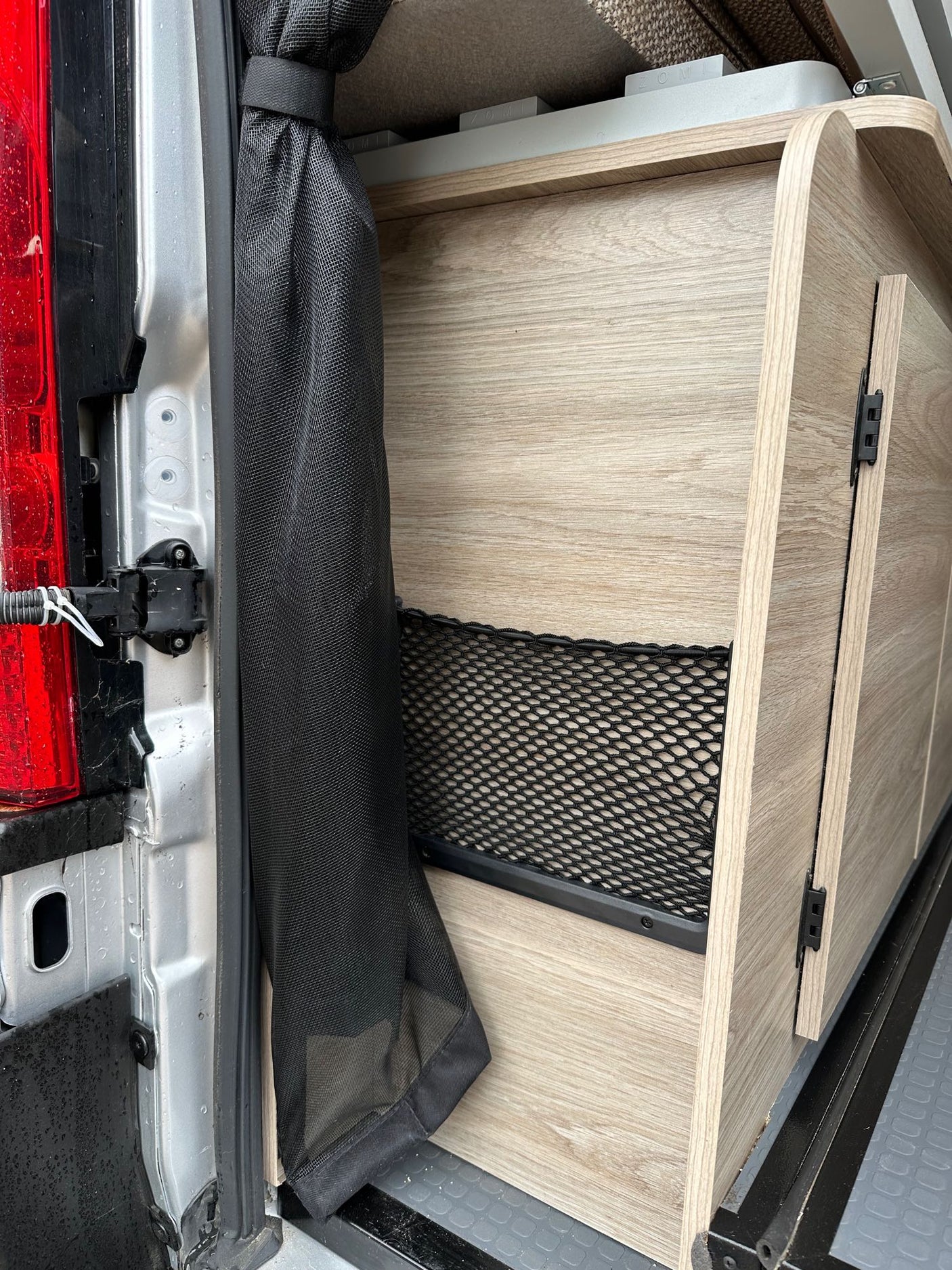 RV rental interior: wood-look storage unit with net pocket & mesh screen near the vehicle's rear door.