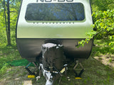 Front view of a "No Boundaries" travel trailer (RV rental) at a wooded campsite, showing tanks & deployed stabilizers.