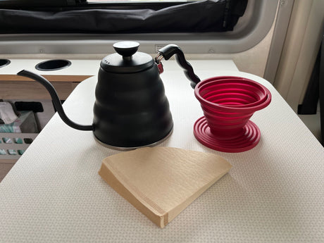 RV rental coffee brewing essentials: A black gooseneck kettle, red collapsible dripper, and filters arranged on a patterned table.