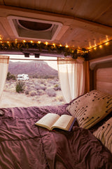 Cozy RV rental bedroom, book on bed, desert view with another RV. Perfect for van life adventure.