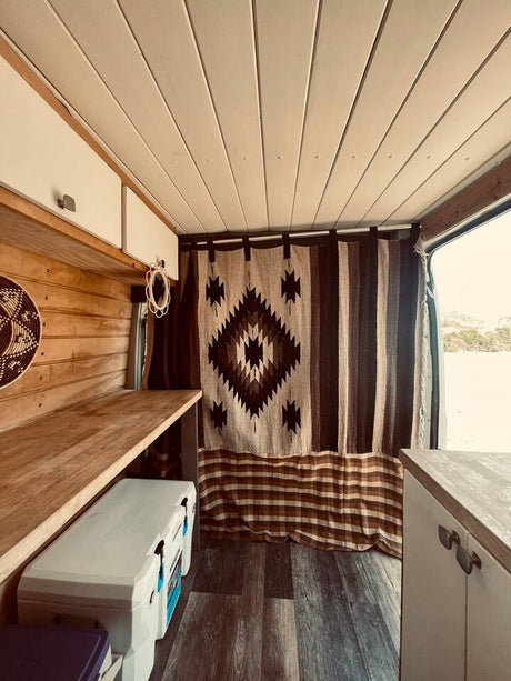 Experience this cozy RV rental's camper interior. Features warm wood paneling, white cabinets, and a unique patterned curtain. Cooler visible.