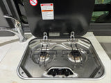 This RV rental kitchen features a compact two-burner gas stovetop with a safety warning on its cover and a chrome faucet.