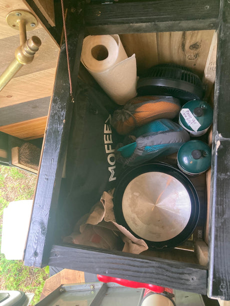 Organized RV rental compartment with camping gear: propane, fan, pan, paper towels, and mesh bags of supplies.