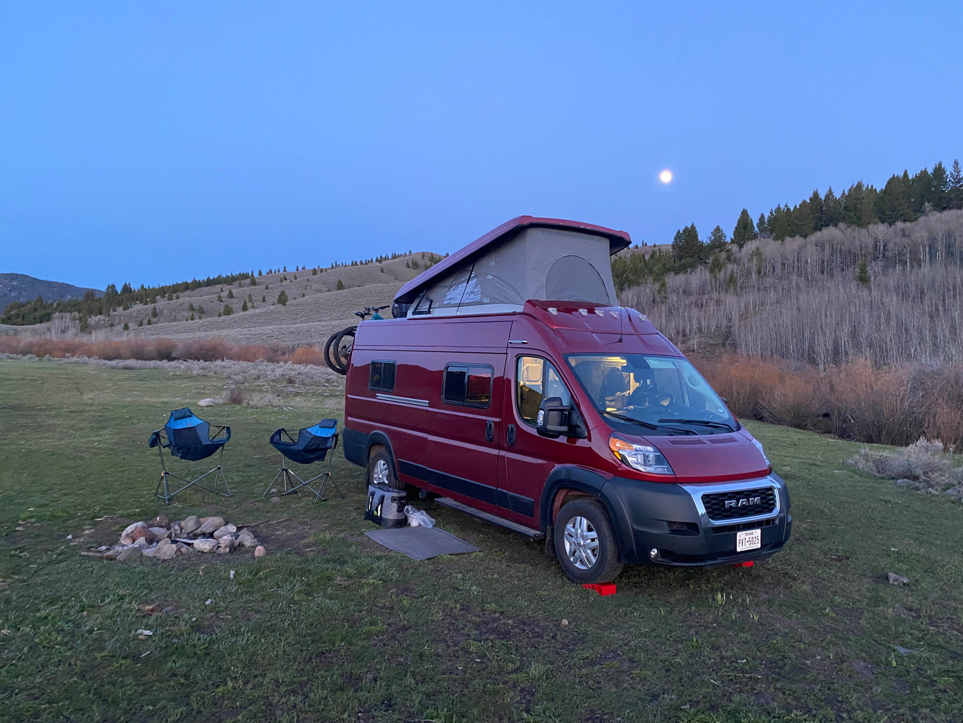 Enjoy the outdoors! This red RV rental camper van, chairs, and fire pit are set for a moonlit camping night.
