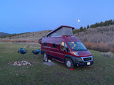 Enjoy the outdoors! This red RV rental camper van, chairs, and fire pit are set for a moonlit camping night.