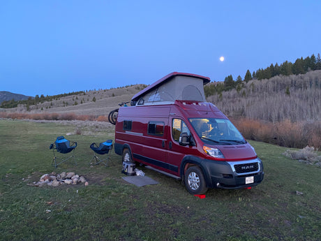 Enjoy the outdoors! This red RV rental camper van, chairs, and fire pit are set for a moonlit camping night.