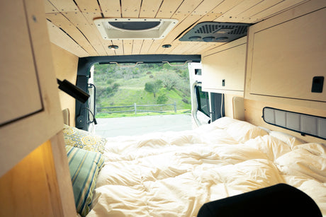 Plush bed inside an RV rental van, with wooden walls & ceiling, overlooks a green hillside. Ready for adventure!