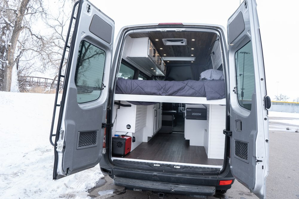 RV rental: Inside a custom camper van with open rear doors, showcasing a raised bed, modern storage, & winter scene.