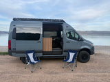 Gray Sprinter **rv rental** van with an open side door, two blue-and-white chairs, parked by a scenic lake and mountains.
