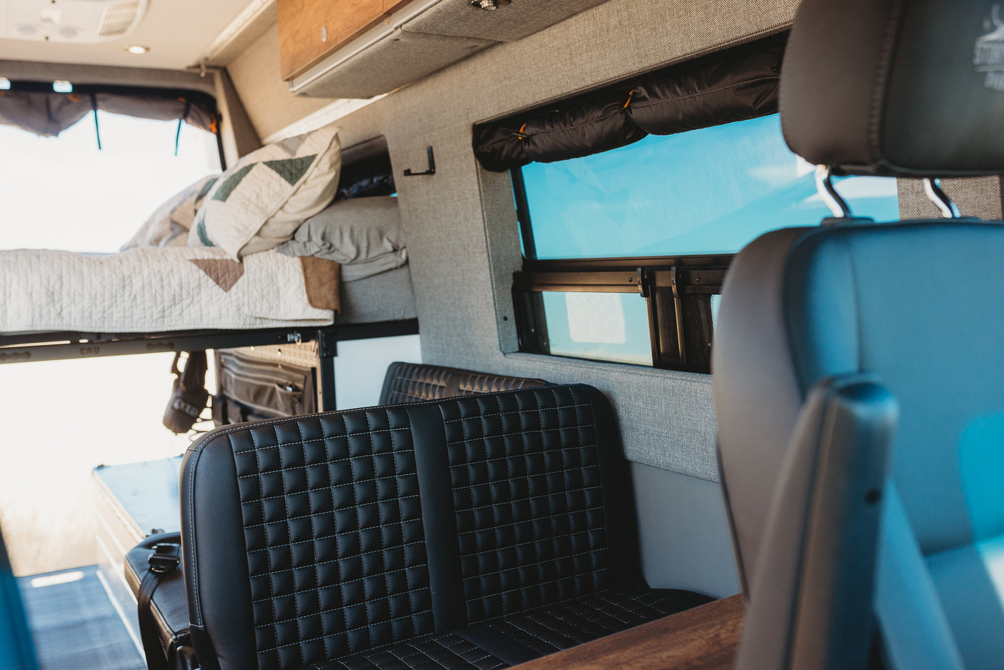 Inside an RV rental: a bright camper van interior with an elevated bed, black quilted seats, and a large window.