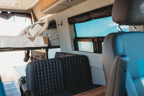 Inside an RV rental: a bright camper van interior with an elevated bed, black quilted seats, and a large window.