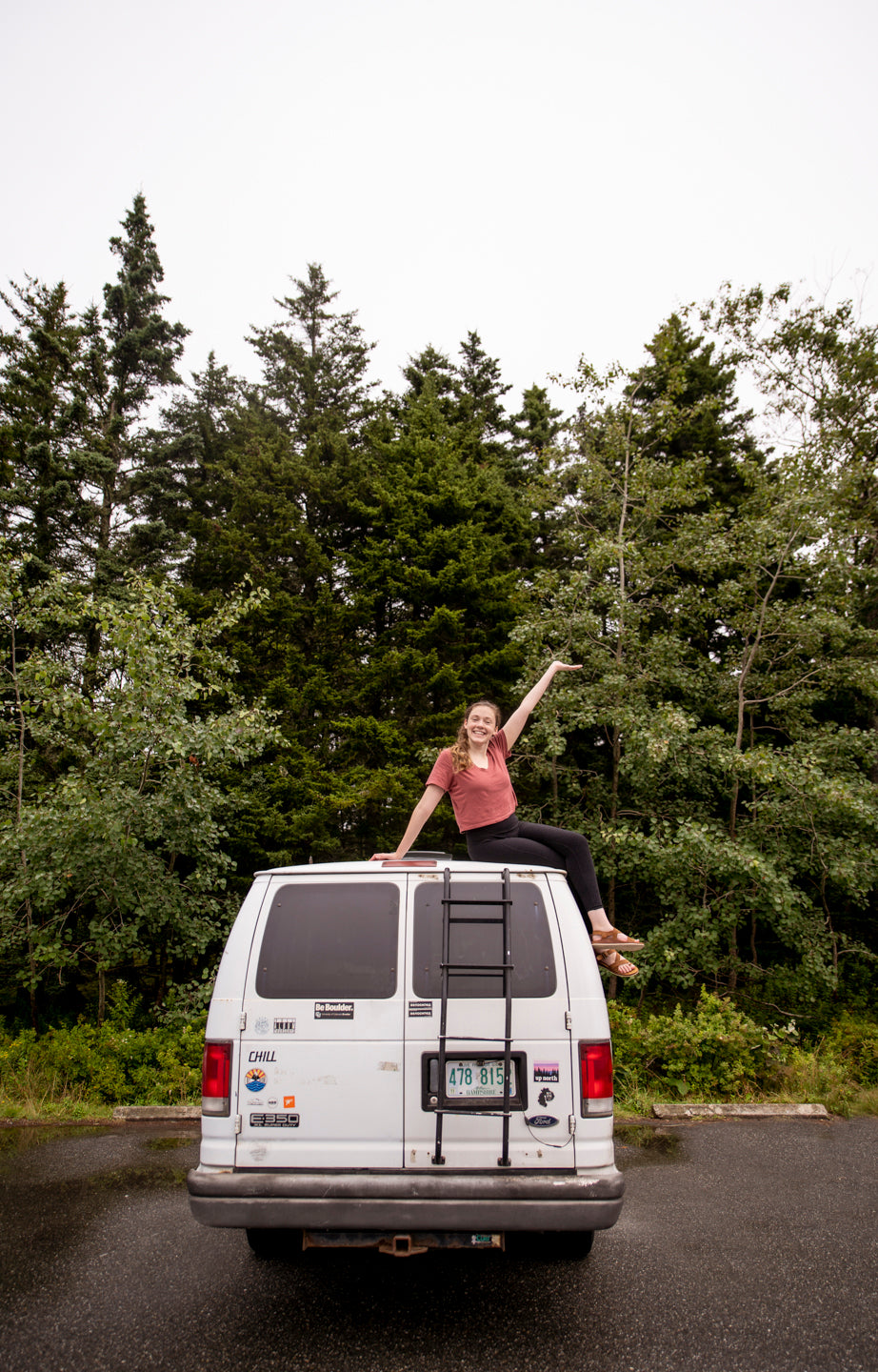 A smiling woman enjoys the view from her white RV rental van's roof, surrounded by tall green forest trees. A peaceful outdoor travel scene.