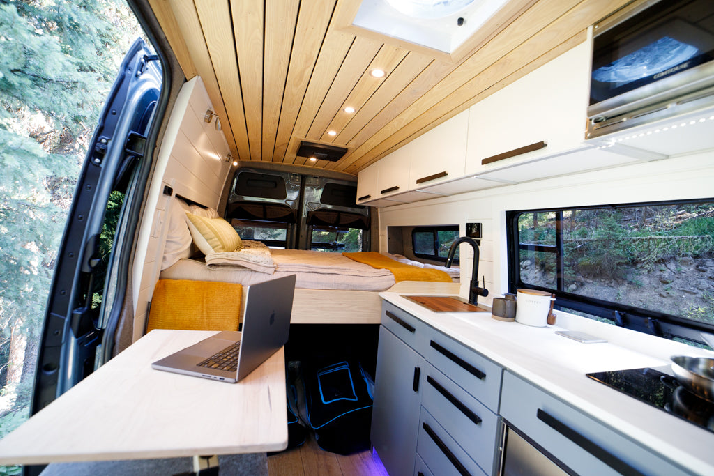 Experience van life! This modern RV rental interior features a cozy bed, full kitchen, and workspace overlooking scenic nature.