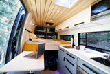 Experience van life! This modern RV rental interior features a cozy bed, full kitchen, and workspace overlooking scenic nature.