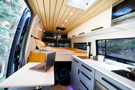 Experience van life! This modern RV rental interior features a cozy bed, full kitchen, and workspace overlooking scenic nature.