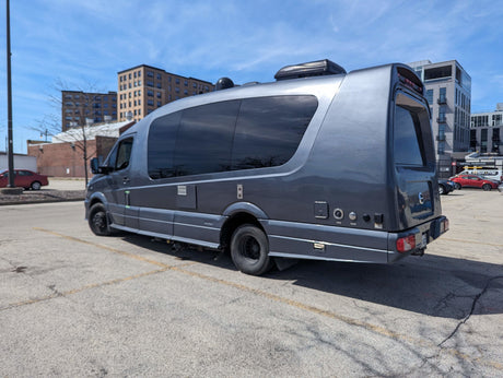Sleek grey RV rental (motorhome) with rooftop AC unit, parked in an urban lot under a sunny sky.