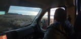 From an **RV rental**, a bearded traveler watches wind turbines, a lake, and sunset over dry plains.