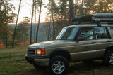 A tan Land Rover with a rooftop tent and awning in a forest, ready for an adventure. Consider this unique rv rental option!