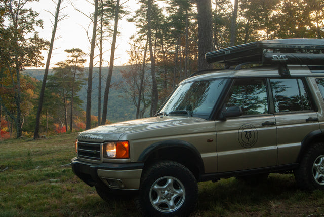 A tan Land Rover with a rooftop tent and awning in a forest, ready for an adventure. Consider this unique rv rental option!
