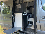 Gray **RV rental** van with open sliding door reveals organized interior: kitchen, controls, passenger seat & pull-out outdoor table.