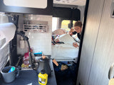 Inside a compact **rv rental**, a boy with a shaka sign peeks from his bed next to a small kitchen area.