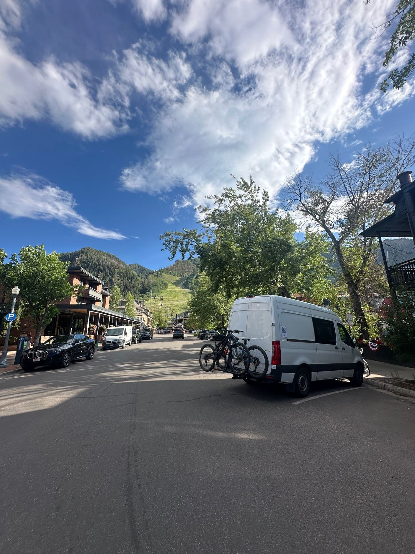 White van with bikes, possibly an RV rental, parked on a sunny street in a vibrant mountain town, ideal for biking adventures.