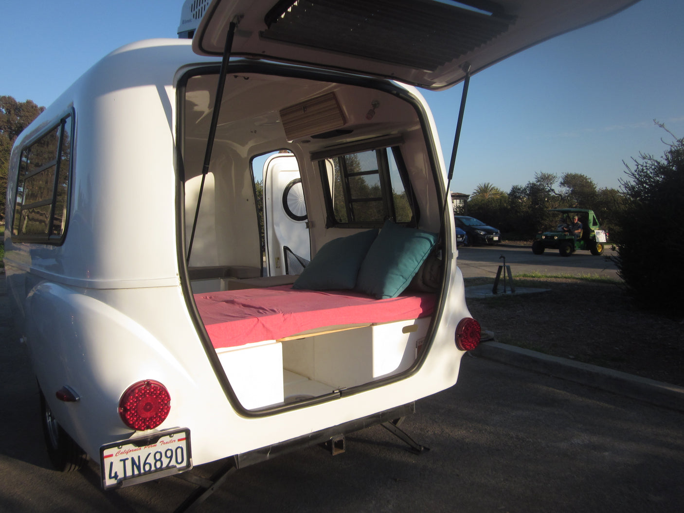 Rear view of a white camper RV rental with its hatch open, revealing a cozy bed with red mattress and teal pillows.