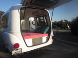 Rear view of a white camper RV rental with its hatch open, revealing a cozy bed with red mattress and teal pillows.