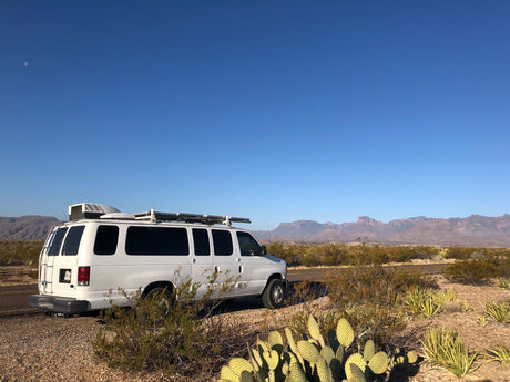 White RV rental van parked roadside in a vast desert landscape. Mountains, clear blue sky with moon, and cacti set a scenic adventure.