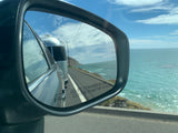 Coastal RV rental adventure: A silver travel trailer is reflected in a side mirror, driving along a scenic ocean road.