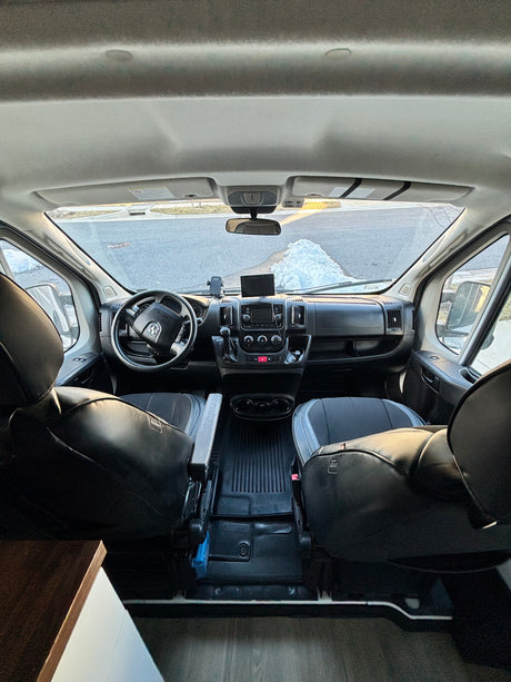 Interior view of an RV rental van's cockpit, featuring the dashboard, steering wheel, and front seats, with a snowy road visible.