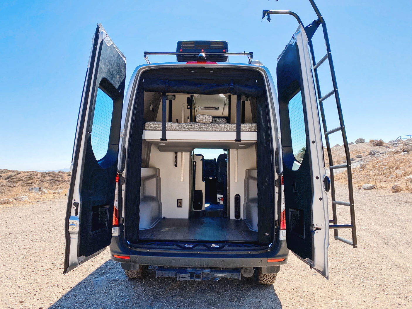 Back of an open RV rental camper van with bunk beds inside, roof rack, and a ladder on the right door. Parked on a dirt road.