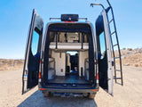 Back of an open RV rental camper van with bunk beds inside, roof rack, and a ladder on the right door. Parked on a dirt road.