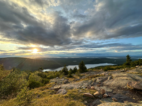 A vibrant sunset casts rays over a vast forested landscape and tranquil lake, ideal for an RV rental getaway.