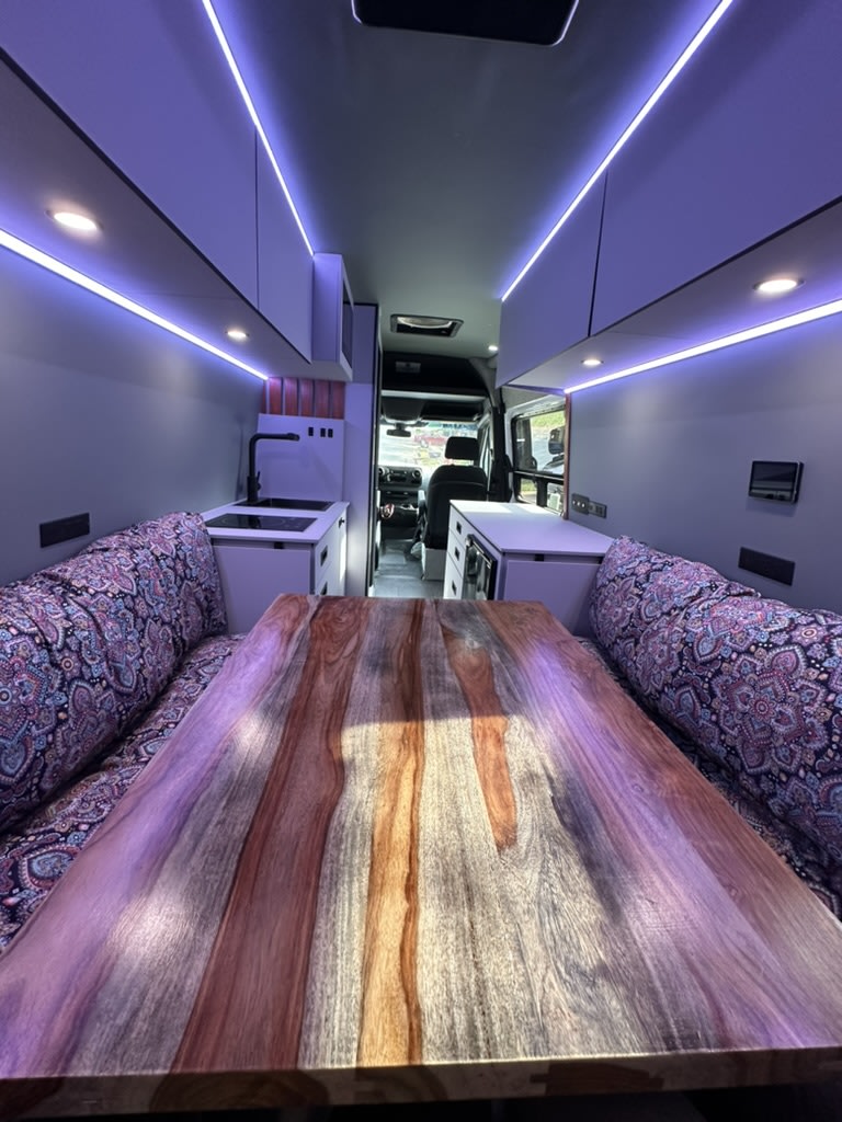  and comfy bench seating.|Woman working on laptop in a cozy **RV rental**. This modern camper van interior is perfect for remote work & travel.|RV rental: Woman slices strawberries on a cutting board in a chic camper van. Bubbly & purple mood lighting enhance the cozy setup.|Productive day in a spacious campervan RV rental! Woman working on her laptop at a wooden table with fruit nearby.|Enjoy a modern RV rental — photo 5