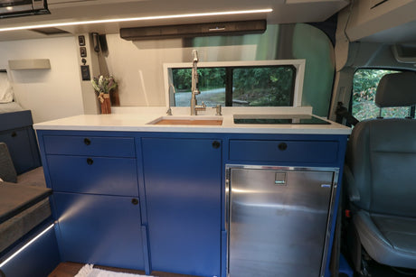 Inside an RV rental: a modern kitchen with blue cabinets, white counters, sink, cooktop, and a view of nature.