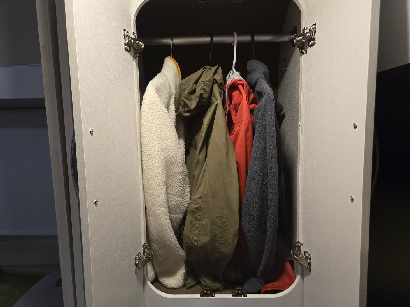 Inside an RV rental: a compact closet with a white fleece, olive jacket, red jacket, and grey fleece hanging.
