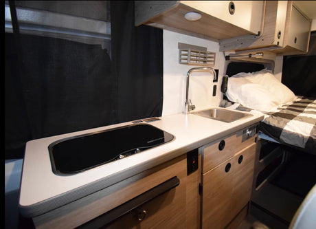 Modern RV rental interior: a functional kitchen with sink, stovetop, and clean countertops, beside a comfy bed.