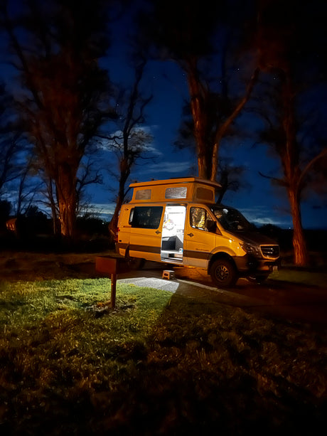 RV rental Sprinter camper van at a night campsite. Its interior glows, pop-top extended, under a starlit, tree-lined sky.