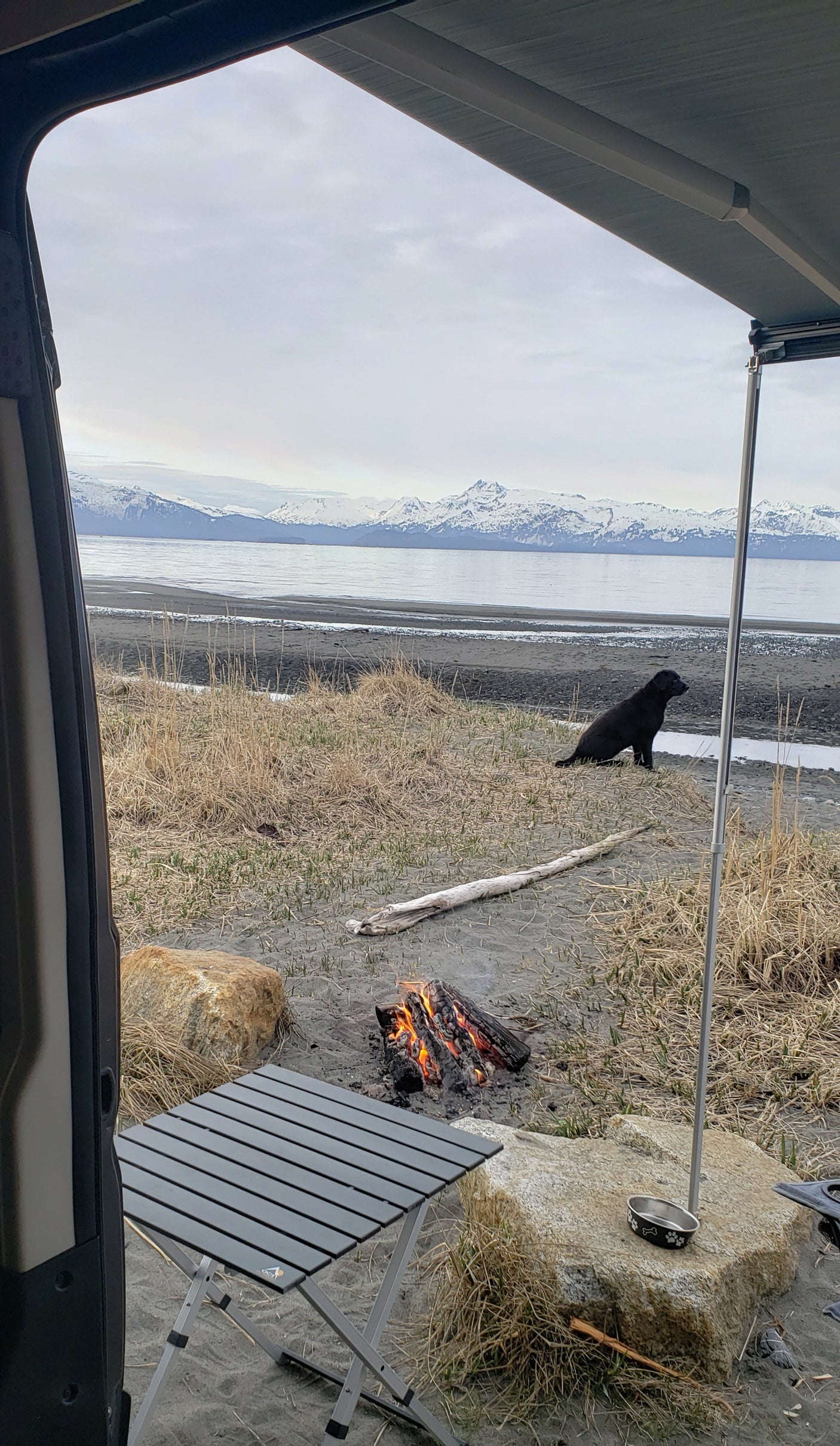 Scenic RV rental view: dog, campfire, and table on a beach with snow-capped mountains across the water.