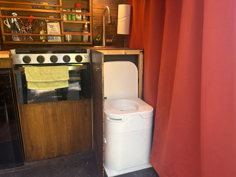 RV rental: Compact kitchen with stove, sink, and shelves next to a portable toilet, partially hidden by a red privacy curtain. — photo 6